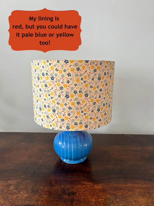 A table lamp with a tiny blue and yellow floral-patterned with red leaves fabric drum shade and a ribbed blue glass base, shown on a dark wooden tabletop against a plain light wall. An orange text bubble at the top reads, 'My lining is red, but you could have it pale blue or yellow too!'.