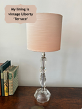 A vintage-style table lamp with a slub dusty pink satin fabric shade and a clear faceted glass base, shown on a wooden table next to books with a text overlay reading 'My lining is vintage Liberty print 'Terrace''.