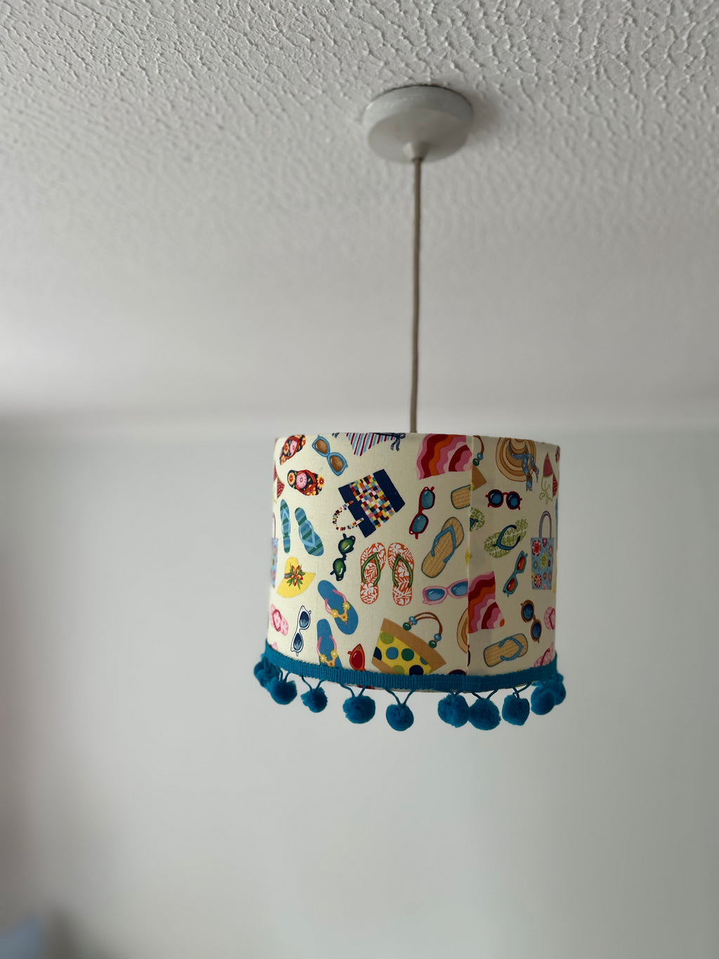 A cylindrical pendant lampshade with a colorful beach-themed print of flip-flops, umbrellas, and sunglasses, finished with a blue pom-pom trim at the bottom, hanging from a ceiling.