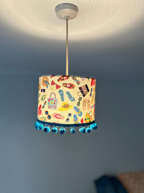 A cylindrical pendant lampshade with a colorful beach-themed print of flip-flops, sunglasses, umbrellas, and towels, finished with a blue pom-pom trim, hanging from a ceiling in a room.
