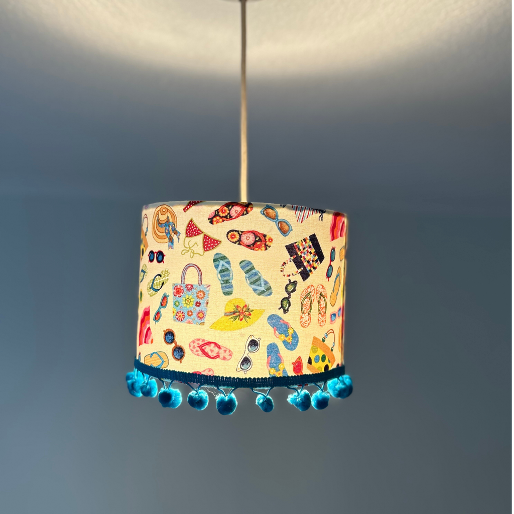A cylindrical pendant lampshade with a colorful beach-themed print of flip-flops, sunglasses, hats, and bags, hanging from a cord against a dark gray background.