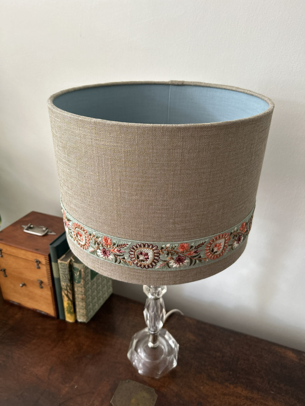 A table lamp with a beige linen drum shade featuring a decorative embroidered band near the bottom, mounted on a clear glass pedestal base. The lamp is set on a dark wooden table next to a small wooden chest and books against a plain off-white wall.
