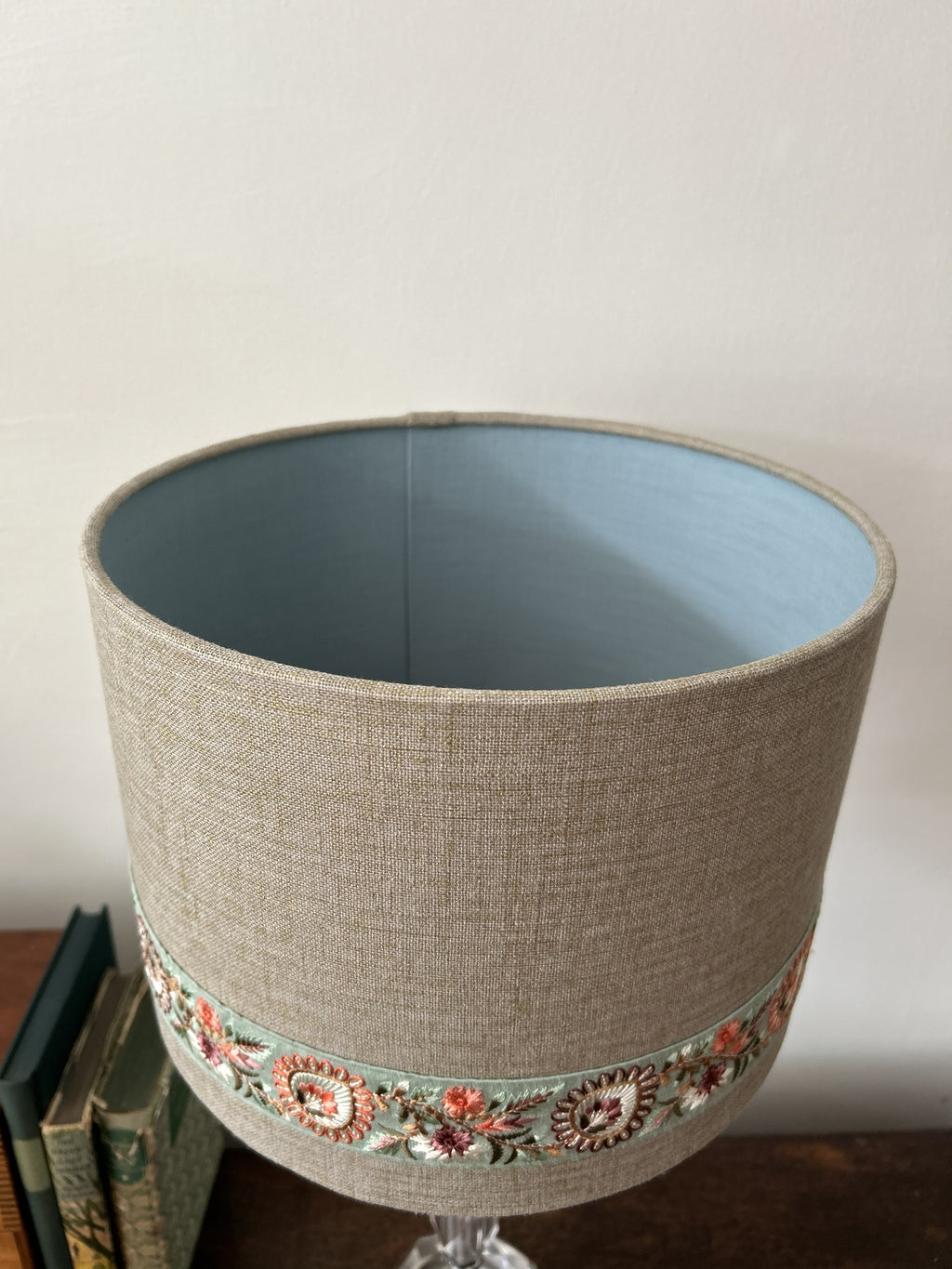 A cylindrical fabric lampshade with a textured beige linen-like exterior, a light blue interior, and a decorative band of colorful floral embroidery near the bottom, shown standing on a table against a plain wall.