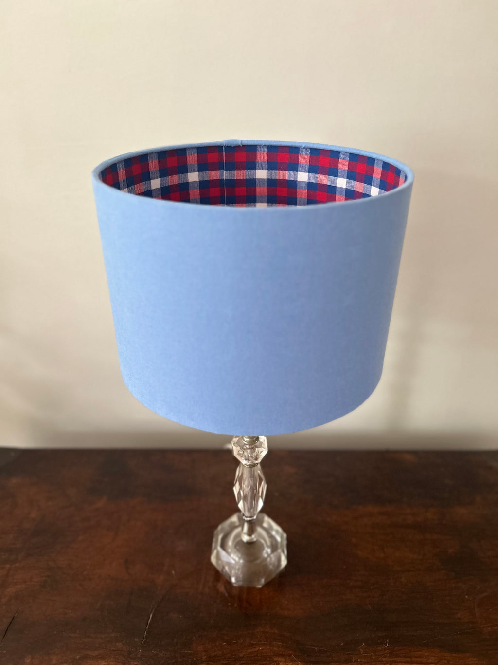 Blue linen drum lampshade on a faceted glass base, with red, navy and white gingham lining visible around the top.