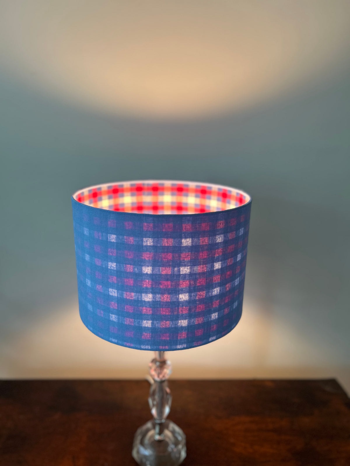 Lit blue linen drum lampshade with red, navy and white gingham checks shining through the outer fabric.