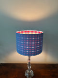 A table lamp with a blue linen outside and a blue and red gingham patterned inside of drum shade and a clear glass stem base, set on a dark wooden surface against a softly lit grey wall.