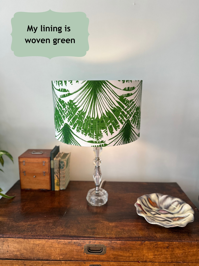 A large table lamp with a white drum shade featuring a green palm leaf pattern, set on a wooden chest. The fabric used for the shade is Brisa' by Michael Szell for Christopher Farr Cloth. A text bubble in the top left reads 'My lining is woven green'.