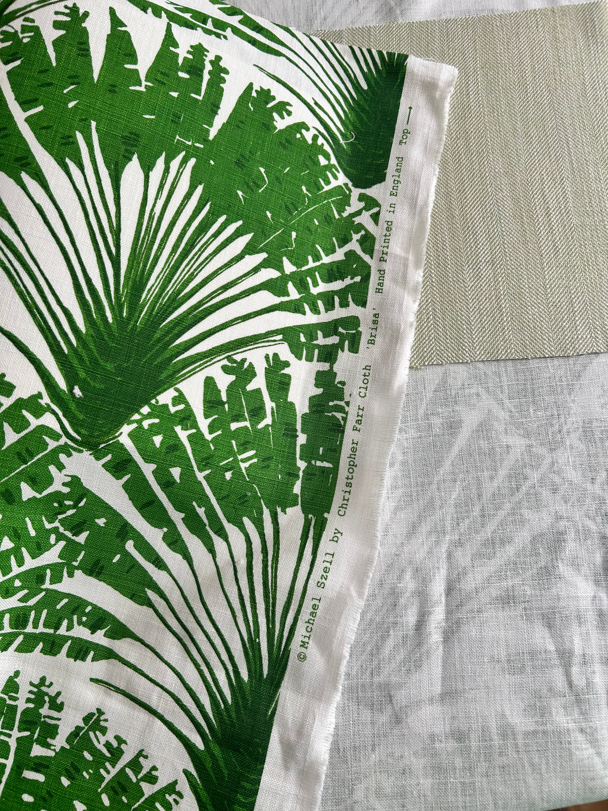 A close-up of a fabric sample featuring a bold green tropical palm leaf pattern on a white background, with a woven beige accent panel and a textured light gray fabric visible behind it.