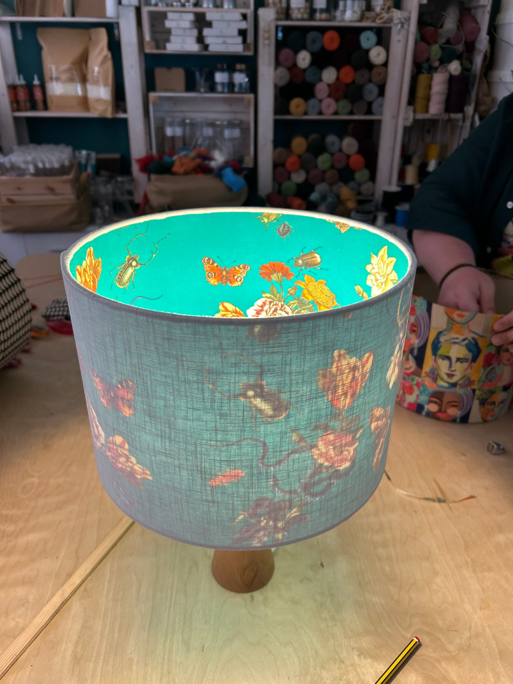 A double sided fabric table lamp shade with a light blue-grey outside and a Victoriana 'cabinet of curiosities' colourful butterfly, beetle, snake and floral print inside, shown on a wooden work bench in a craft room setting.