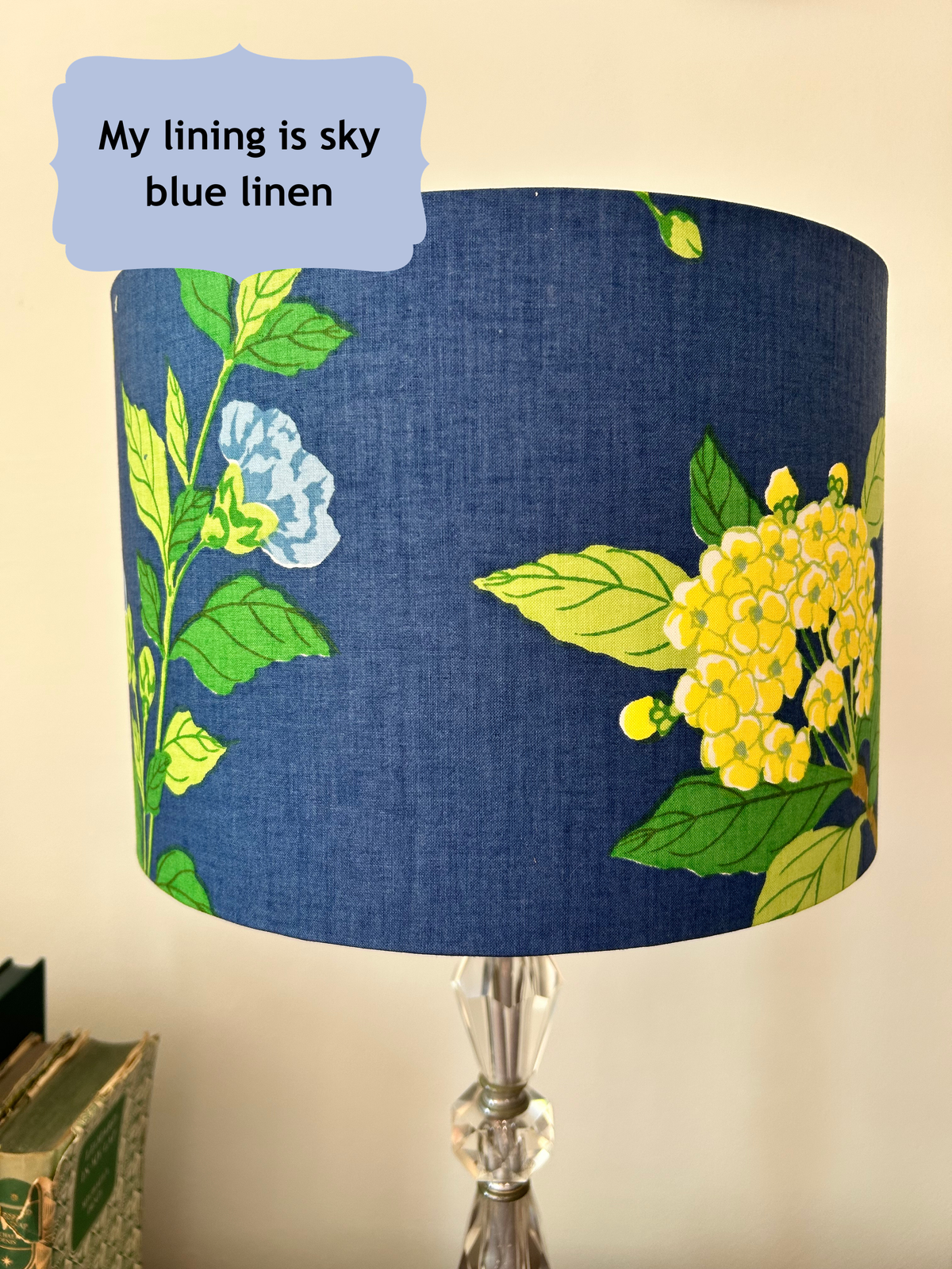 A table lamp shade made of blue linen fabric with a floral print of yellow flowers and green leaves, shown on a lamp stand against a beige wall. A text bubble in the top left reads 'My lining is sky blue linen'.