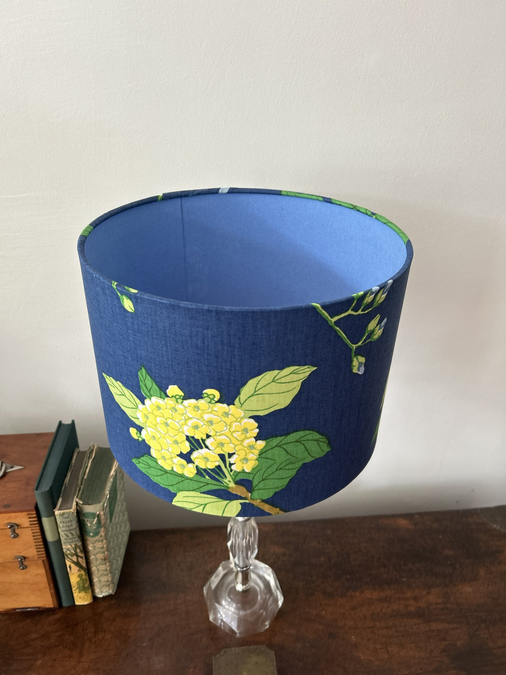 A blue fabric table lampshade with a yellow and green floral print, with a blue fabric inside, mounted on a clear glass stem base and set on a wooden table next to books against a plain wall.