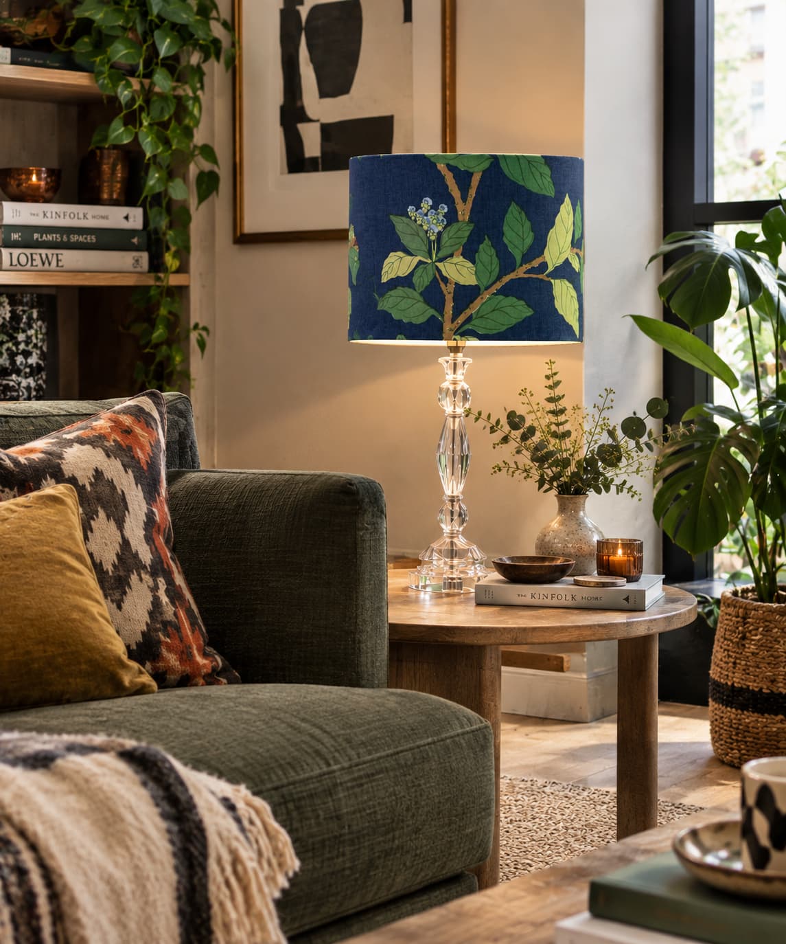 Lifestyle Image of blue, green and yellow fabric drum lampshade. Pattern on the shade is a blue background and Chinese Hydrangeas as the floral pattern. The Shade it lit and the pale blue linen lining can be seen slightly. The shade is on a glass base and in a lounge setting complete with sofa and cushions in natural tones of green, beige, mustard and terracotta. There are numerous house plants, books and candles around the room.