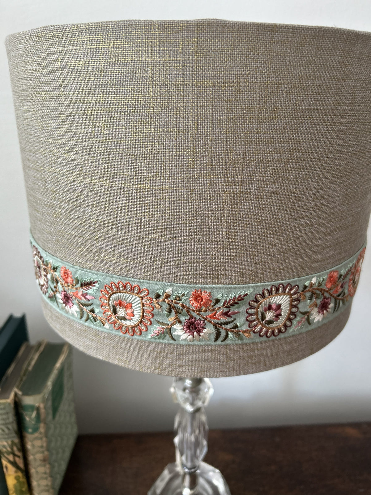 A close-up of a cylindrical table lampshade made of textured beige linen fabric, featuring a decorative band of colorful floral embroidery in pink, orange, and green on a light blue base. The lampshade is mounted on a clear glass stem lamp base, set against a neutral background with books on a wooden surface.