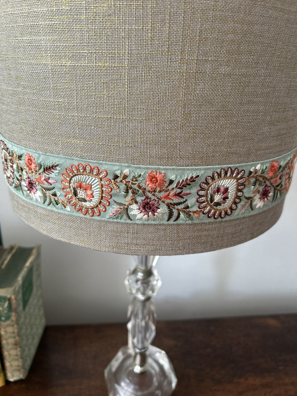 A close-up of a table lamp shade made of textured beige linen, decorated with a wide band of floral embroidery in pink, orange, and green on a mint background, mounted on a clear glass lamp base.