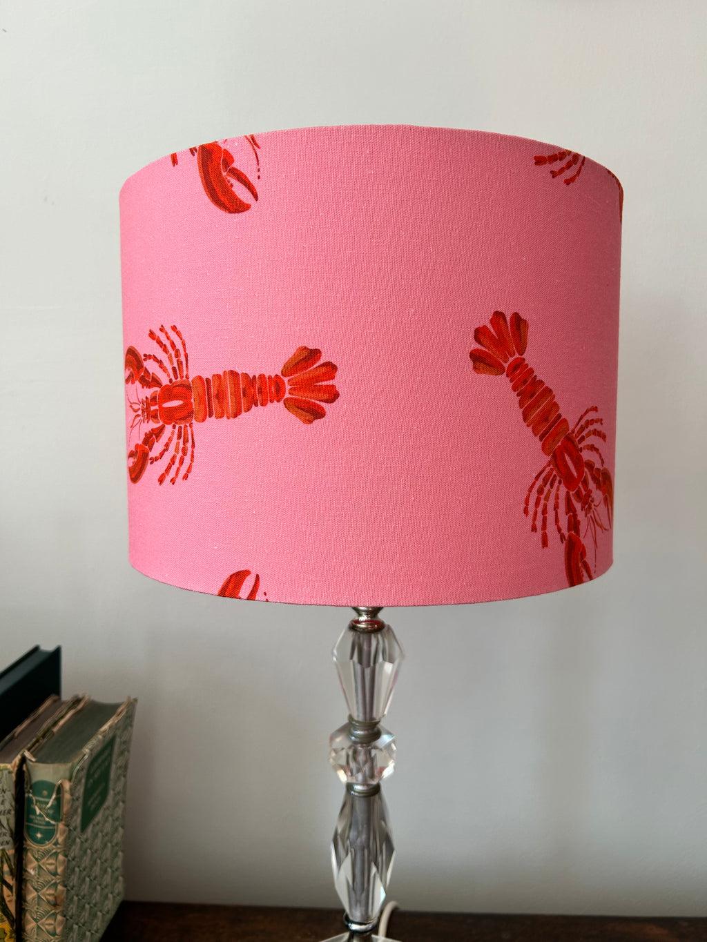 A pink table lamp shade with a red lobster pattern, mounted on a clear faceted glass stem, set against a plain light gray wall with books partially visible on the left.