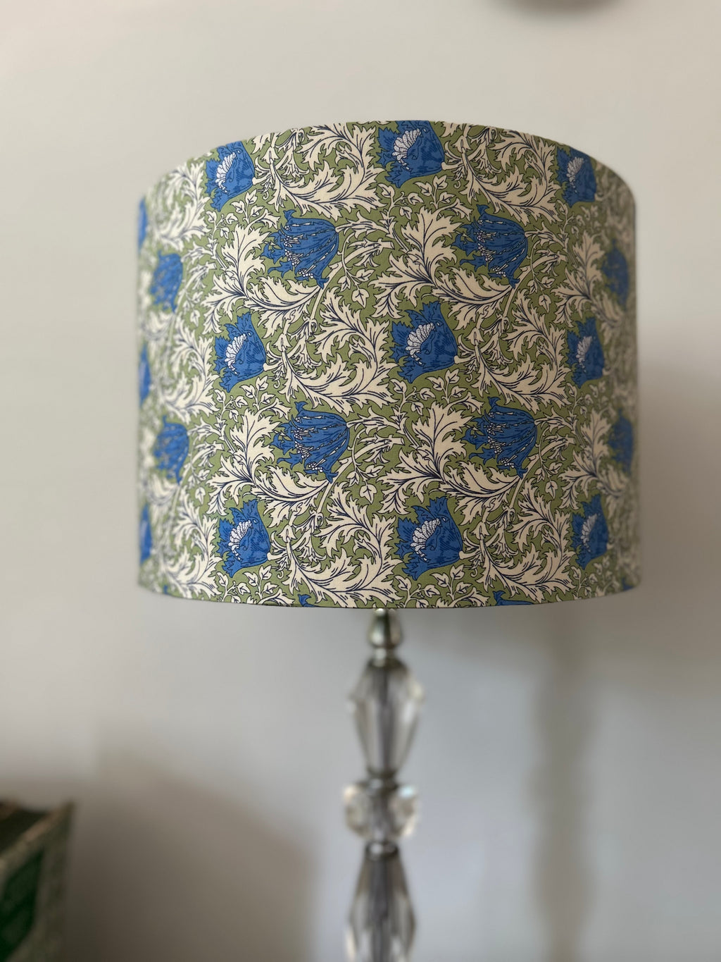 A table lamp with a drum shade featuring a blue and green botanical pattern on a light background, mounted on a clear faceted glass stand against a plain wall.