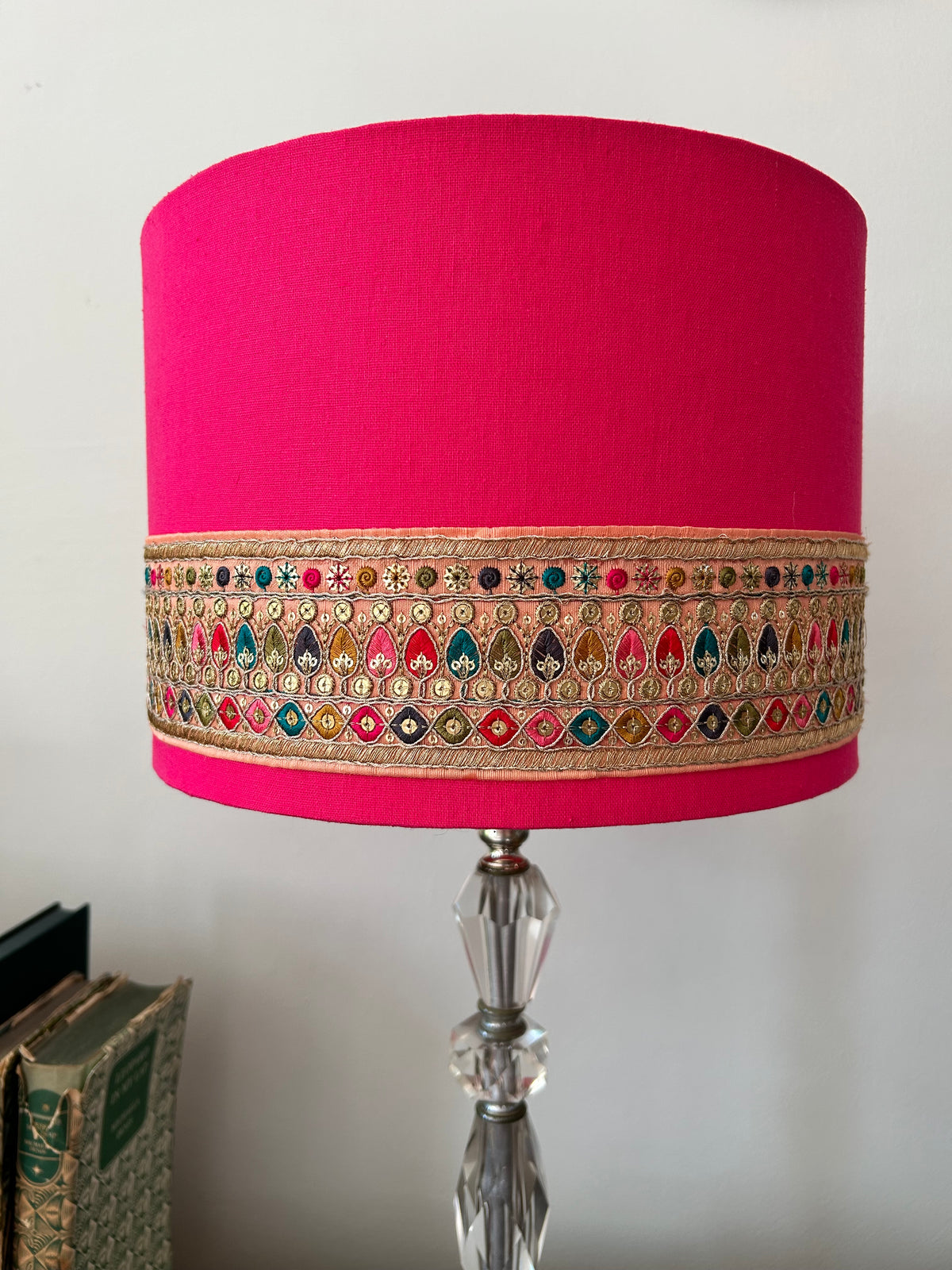 A vibrant hot pink fabric lampshade adorned with a detailed multicolor embroidered band, mounted on a clear faceted glass stand.