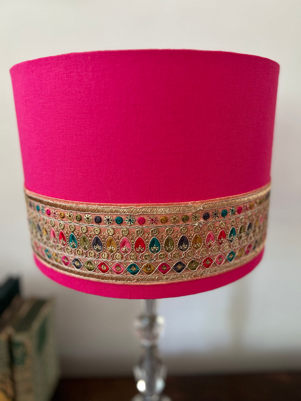 A bright pink fabric lampshade with an ornate gold embroidered band featuring colorful teardrop motifs, shown on a clear glass lamp stand against a neutral indoor background.