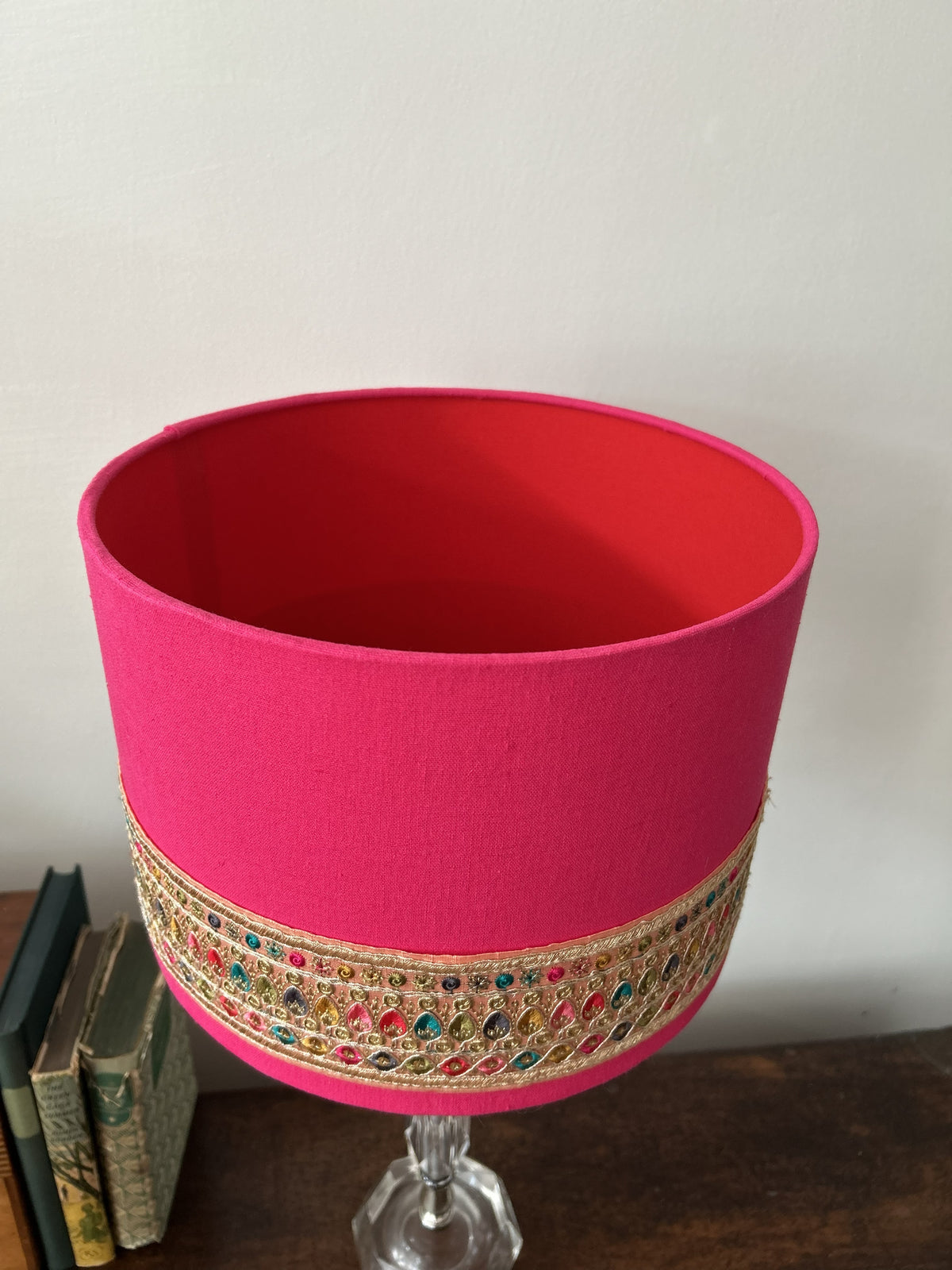 A bright pink cylindrical lampshade with an ornate gold embroidered band featuring colorful rhinestones, displayed on a glass stem holder on a wooden surface.