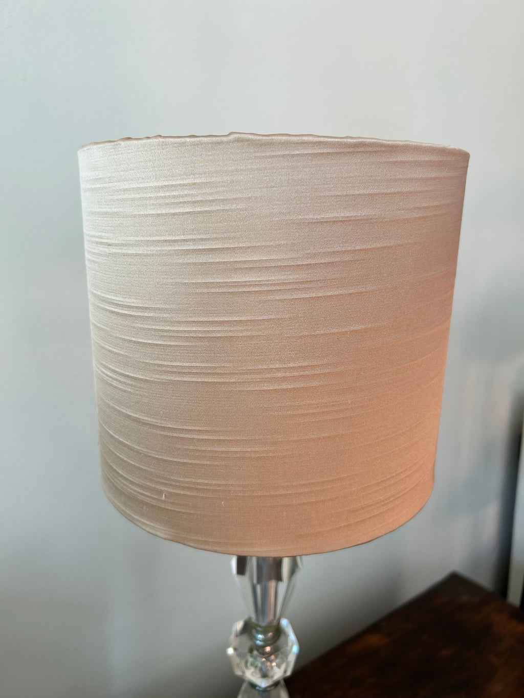 A cylindrical fabric lampshade in a warm beige or light tan colour with slub satin texture, mounted on a clear faceted glass and silver metal lamp base, set against a neutral gray wall.