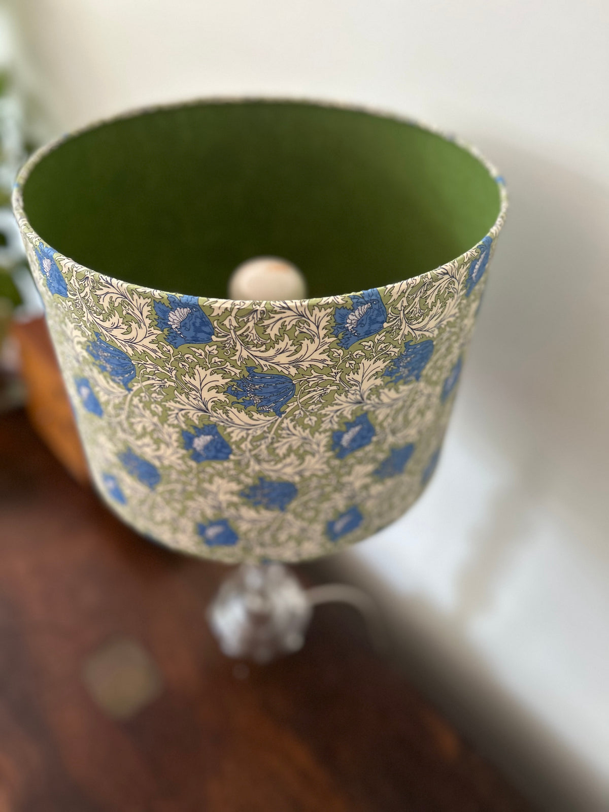 A decorative lampshade with a blue and green floral pattern on the exterior and a solid olive green interior, shown resting on a dark wooden surface.