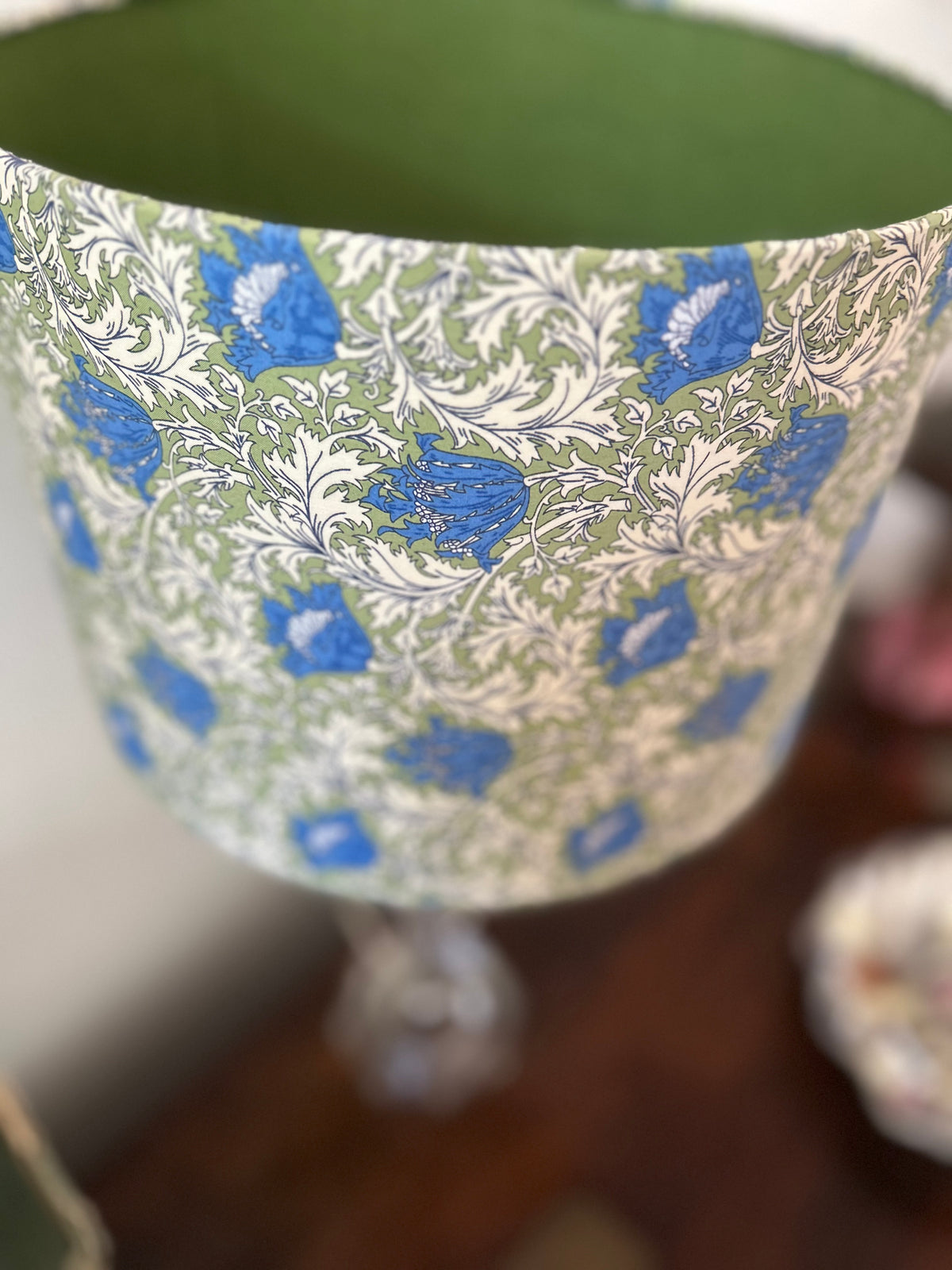 A close-up of a lampshade with a green and blue floral pattern, featuring a solid green inner lining, set against a blurred background.