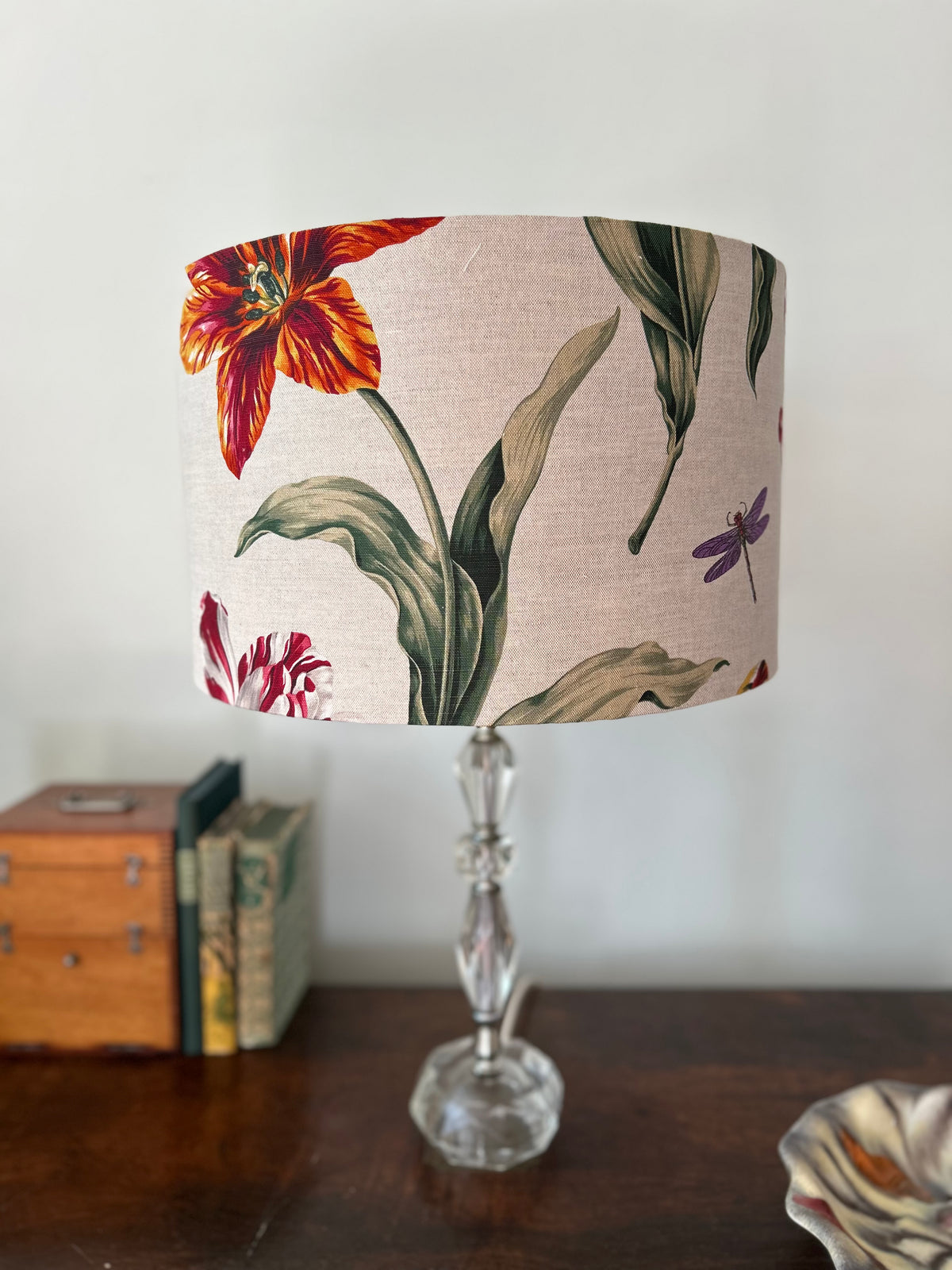 A table lamp with a beige linen drum shade featuring a botanical print of red and orange tulips, green leaves, and a purple dragonfly, set on a dark wooden table against a white wall.