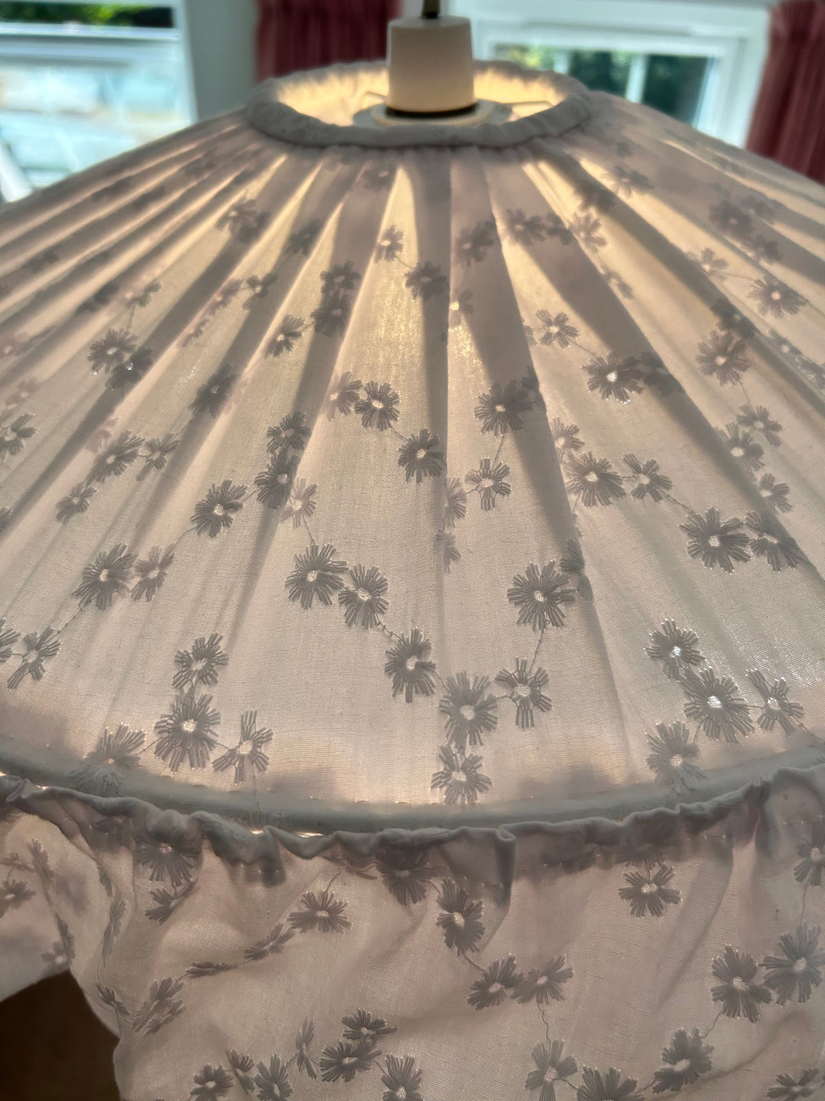 angled view of white broderie anglaise romantic, cottage core, frilly ceiling lampshade which is lit. The glow of the light is coming through the fabric highlighting the pleats, folds and frills.