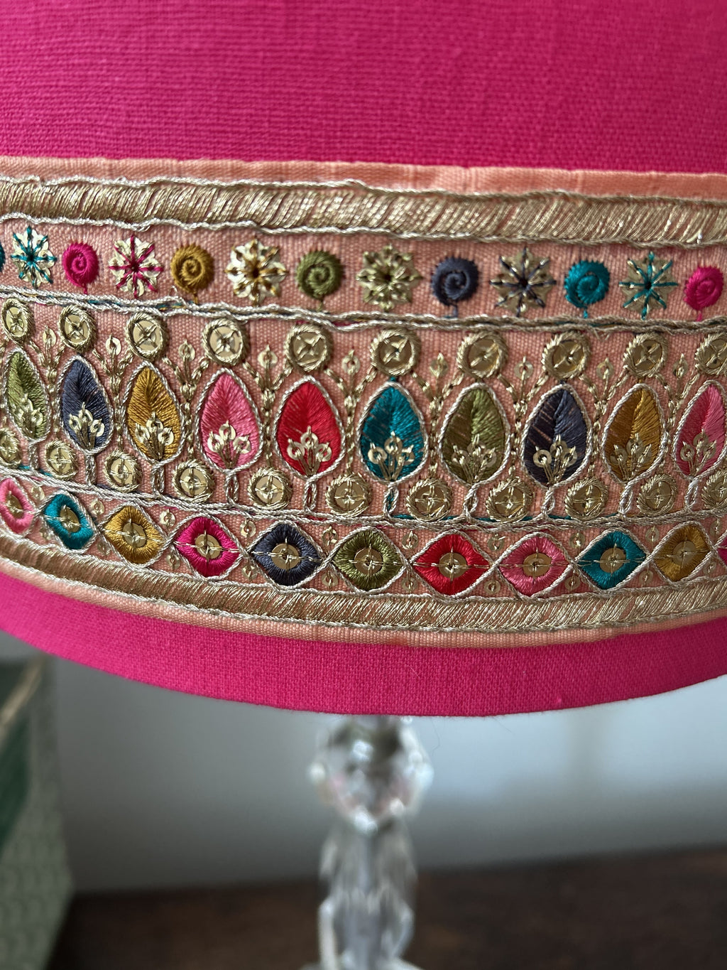 Close-up of a pink fabric lampshade with ornate multicolor embroidery and gold metallic detailing around the lower edge, mounted on a clear glass lamp stand.