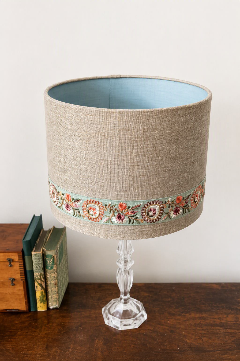 A table lamp with a beige linen drum shade featuring a colorful floral embroidered band near the bottom, a light blue interior lining, and a clear faceted glass pedestal base, shown on a wooden surface next to stacked books against a plain wall.