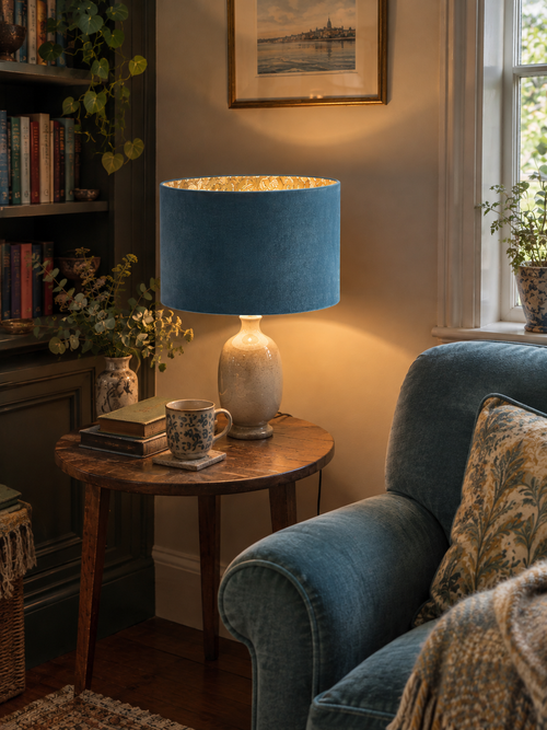  Blue velvet lampshade with glowing golden floral lining beside a blue armchair and bookcase.