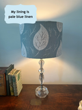 A table lamp with a scandi vibe blue fabric shade featuring a white leaf pattern, set on a wooden table against a blue wall. A text bubble in the top left reads 'My lining is pale blue linen'.