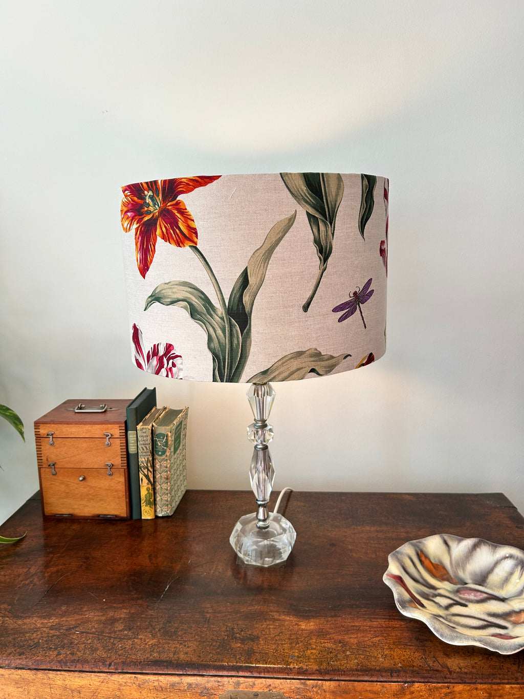 A table lamp with a beige fabric drum shade featuring a botanical print of red flowers, green leaves, and purple dragonflies, set on a dark wooden table next to books and a decorative bowl.