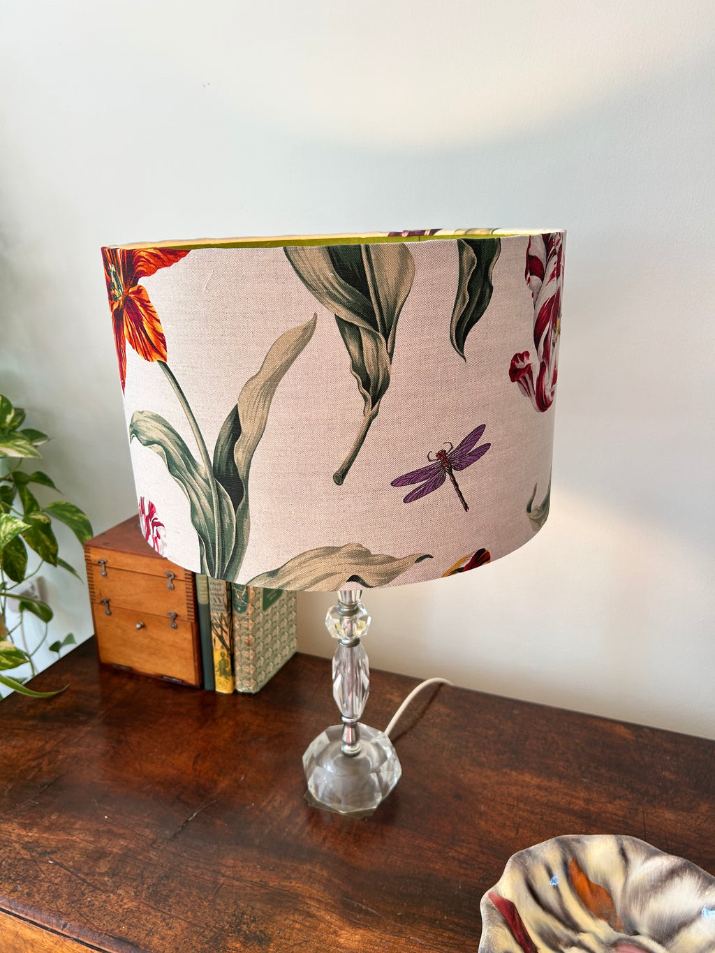 A table lamp with a beige fabric drum shade featuring a botanical and dragonfly pattern, set on a dark wooden table next to a small wooden box and a potted plant.