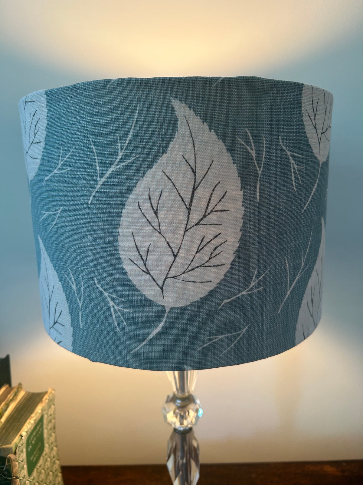 Close front view of a lit duck-egg blue drum lampshade, showing a large white leaf with dark veins and fine twig motifs.