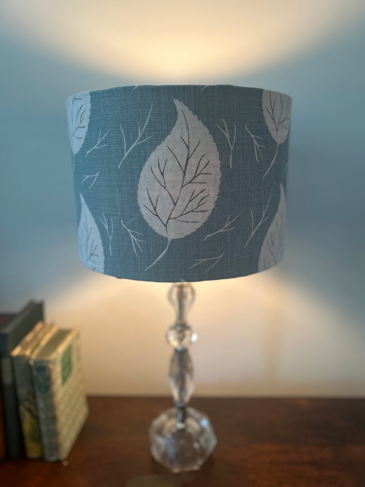 Lit duck-egg blue drum lampshade with white leaves and fine twig motifs, displayed on a clear glass base beside books.