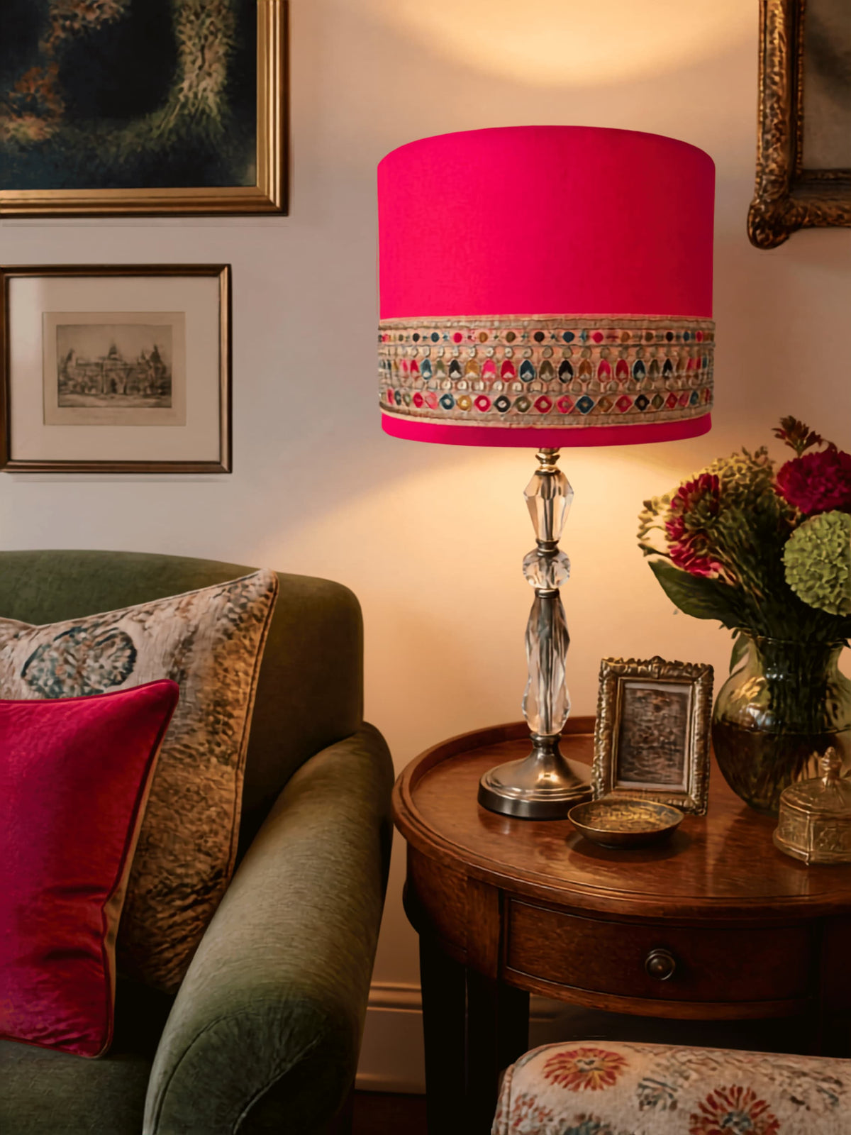 A vibrant hot pink table lamp with a patterned embroidered band around the lower edge, set on a wooden side table next to a green armchair in a cozy living room.