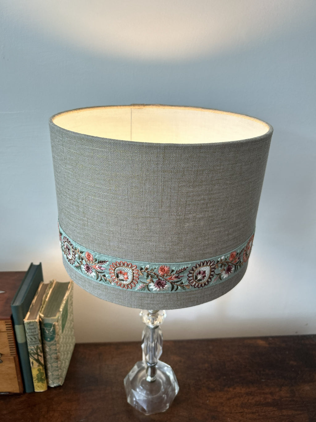 A table lamp with a cylindrical gray linen shade featuring a decorative embroidered band and a clear faceted glass base, shown on a wooden surface against a light wall.