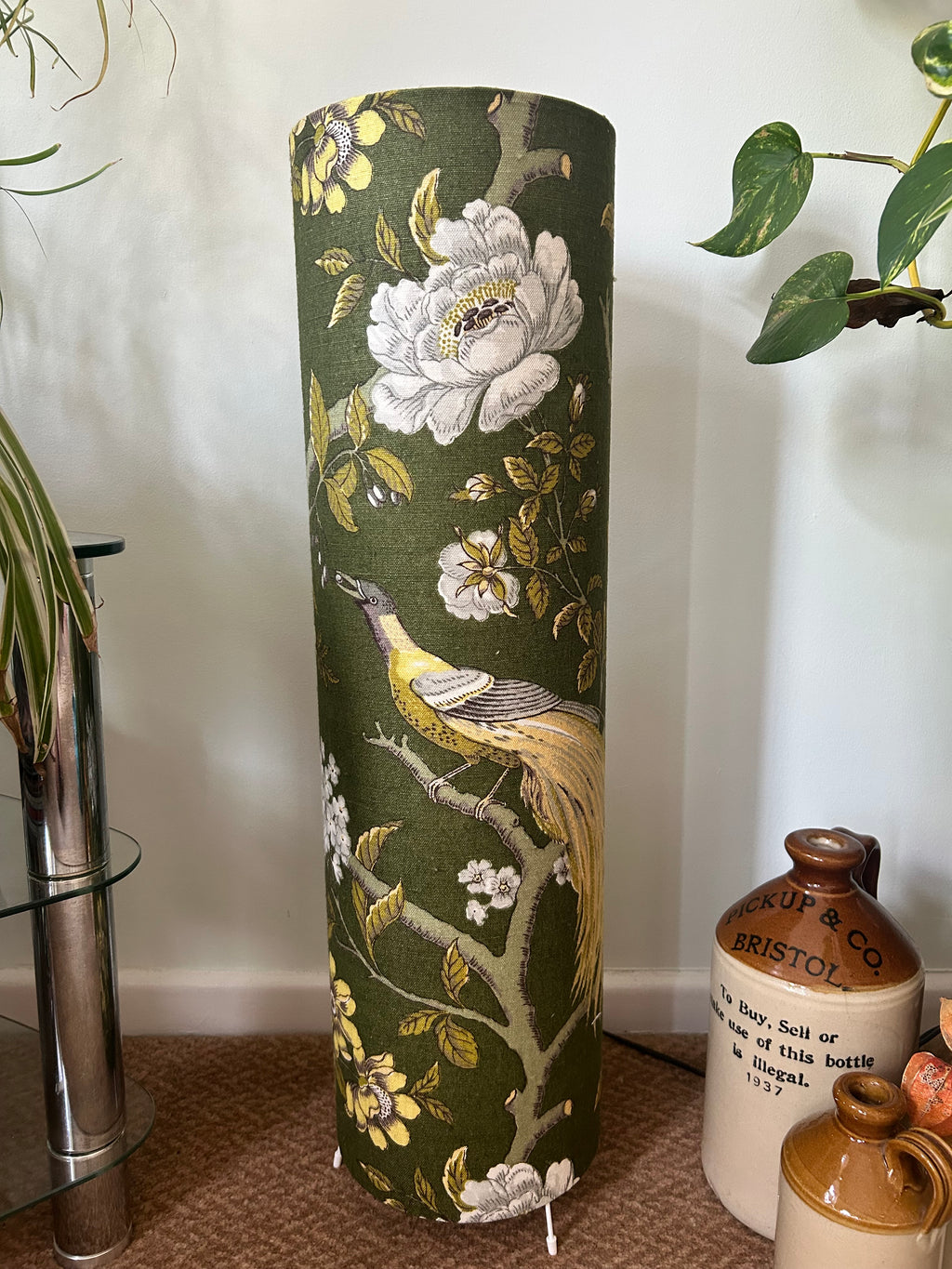 A tall cylindrical floor lamp covered in dark olive green fabric with a classic floral and bird print, standing on a carpeted floor in a home setting.