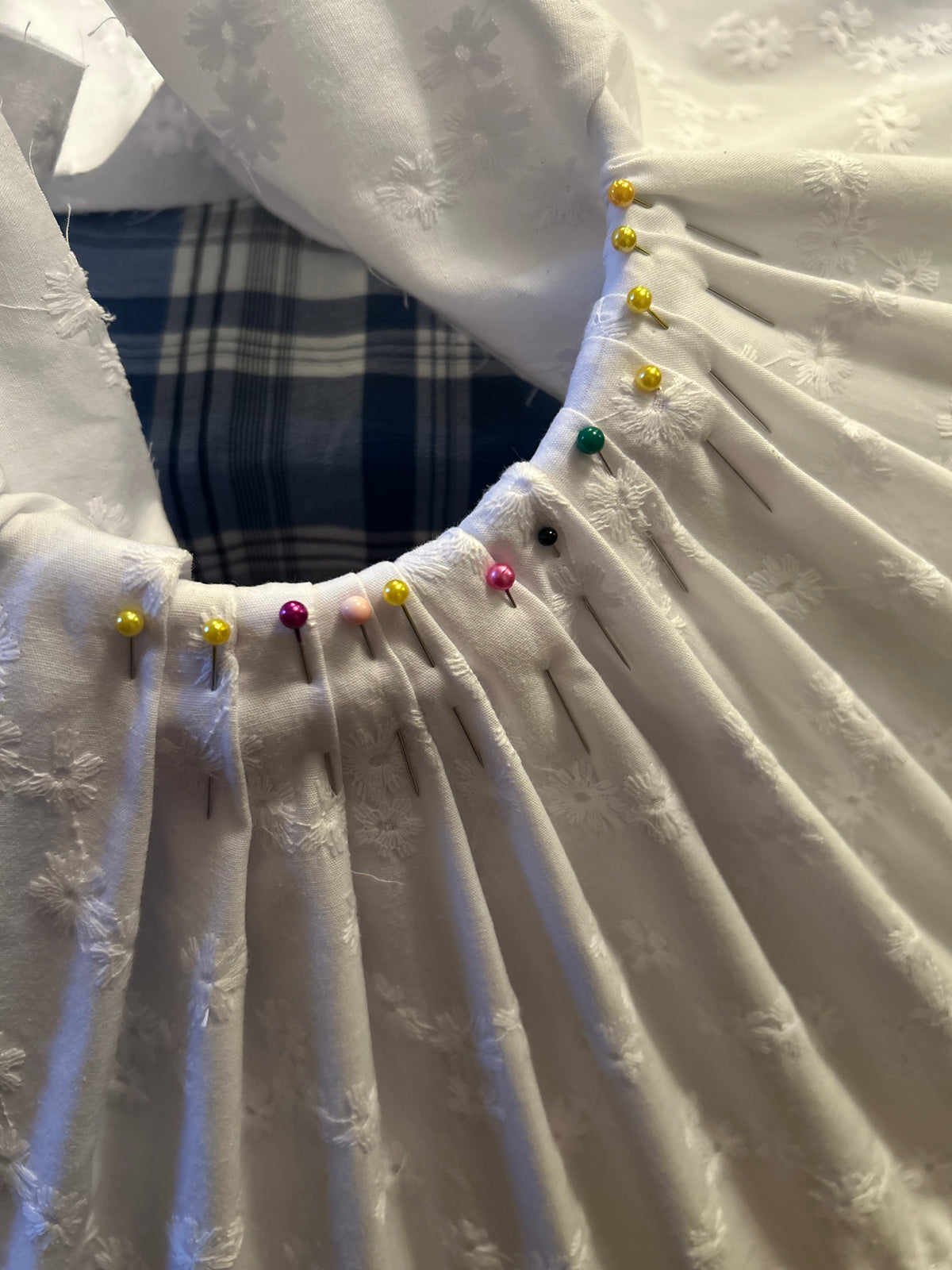 Close-up of a white broderie anglaise fabric shade with gathered pleats and colourful sewing pins along the top opening of the shade, with a plaid fabric visible underneath.