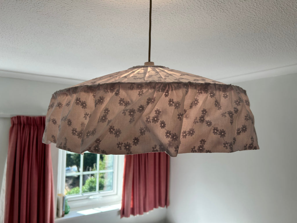 A vintage-style cottage core pendant lampshade made with broderie anglaise white fabric with a floral pattern, hanging from a cord in a room with a window and pink curtains. The light is on and there's a soft glow through the cotton fabric.