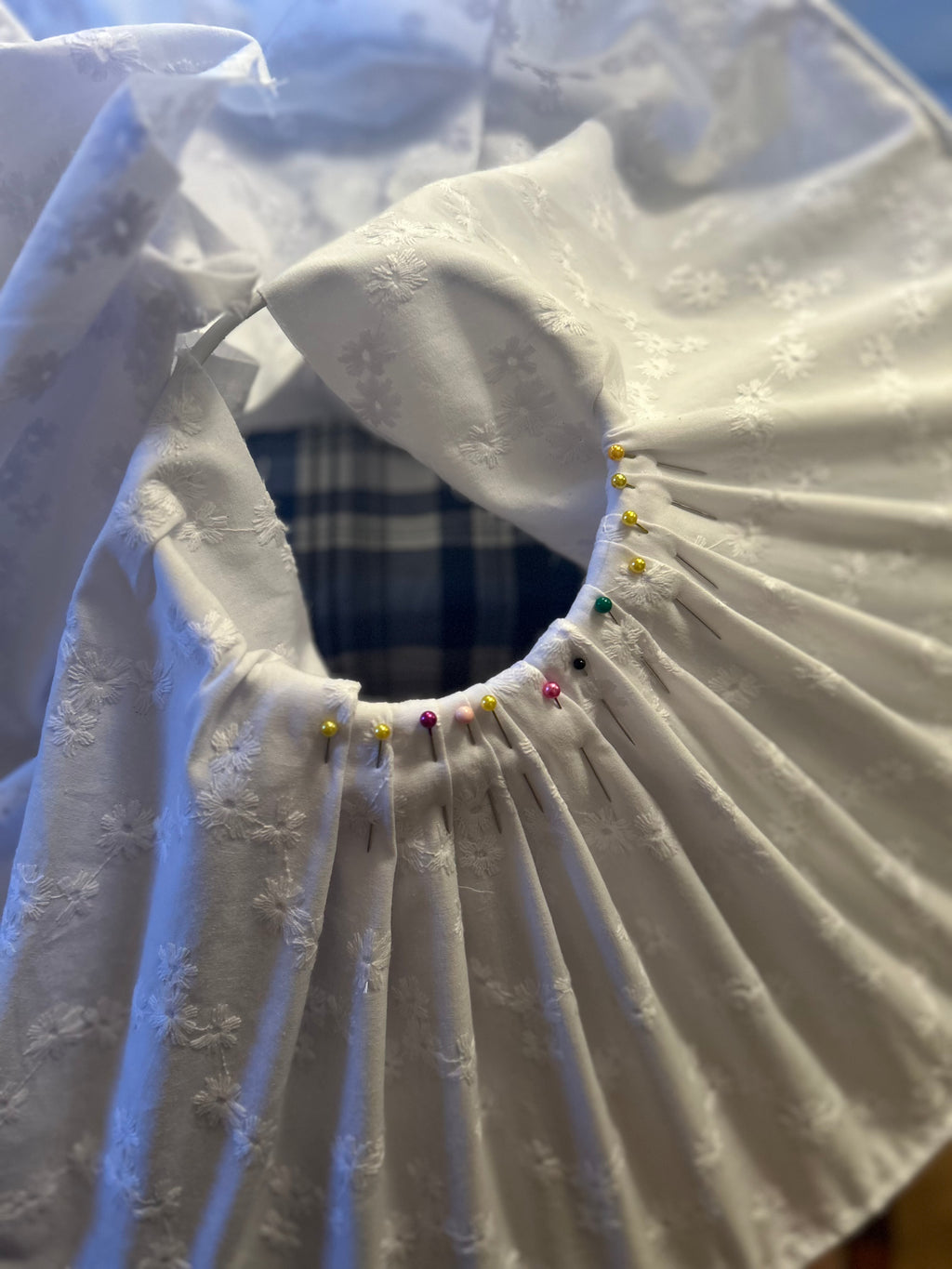 A close-up of a white textured broderie anglaise fabric shade with gathered pleats around a neckline, part-secured with colourful sewing pins, on a blue plaid surface.