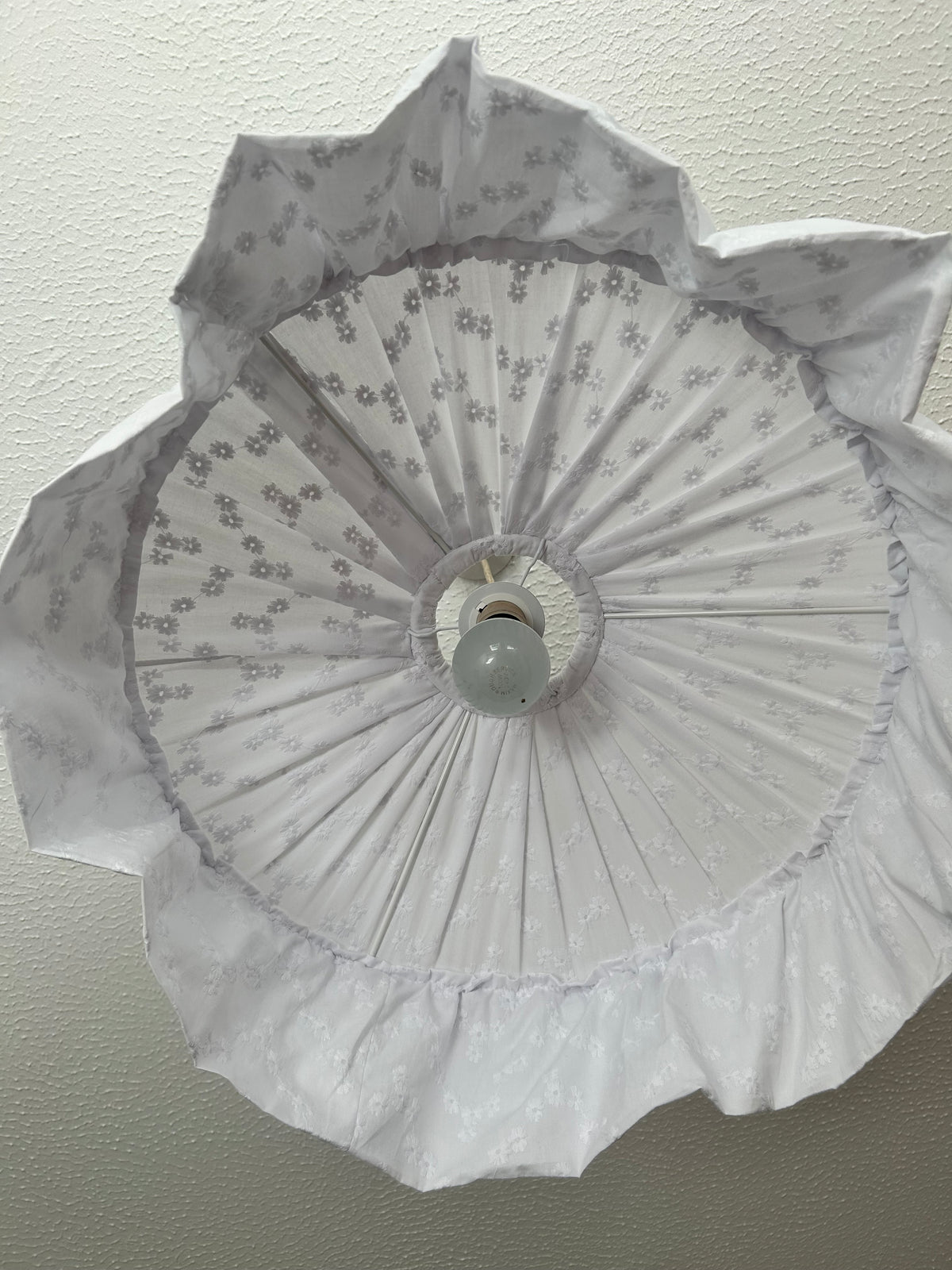 A white broderie anglaise fabric flower-large circular shaped ceiling light fixture with a central bulb socket and a small opaque bulb, shown against a textured off-white ceiling.