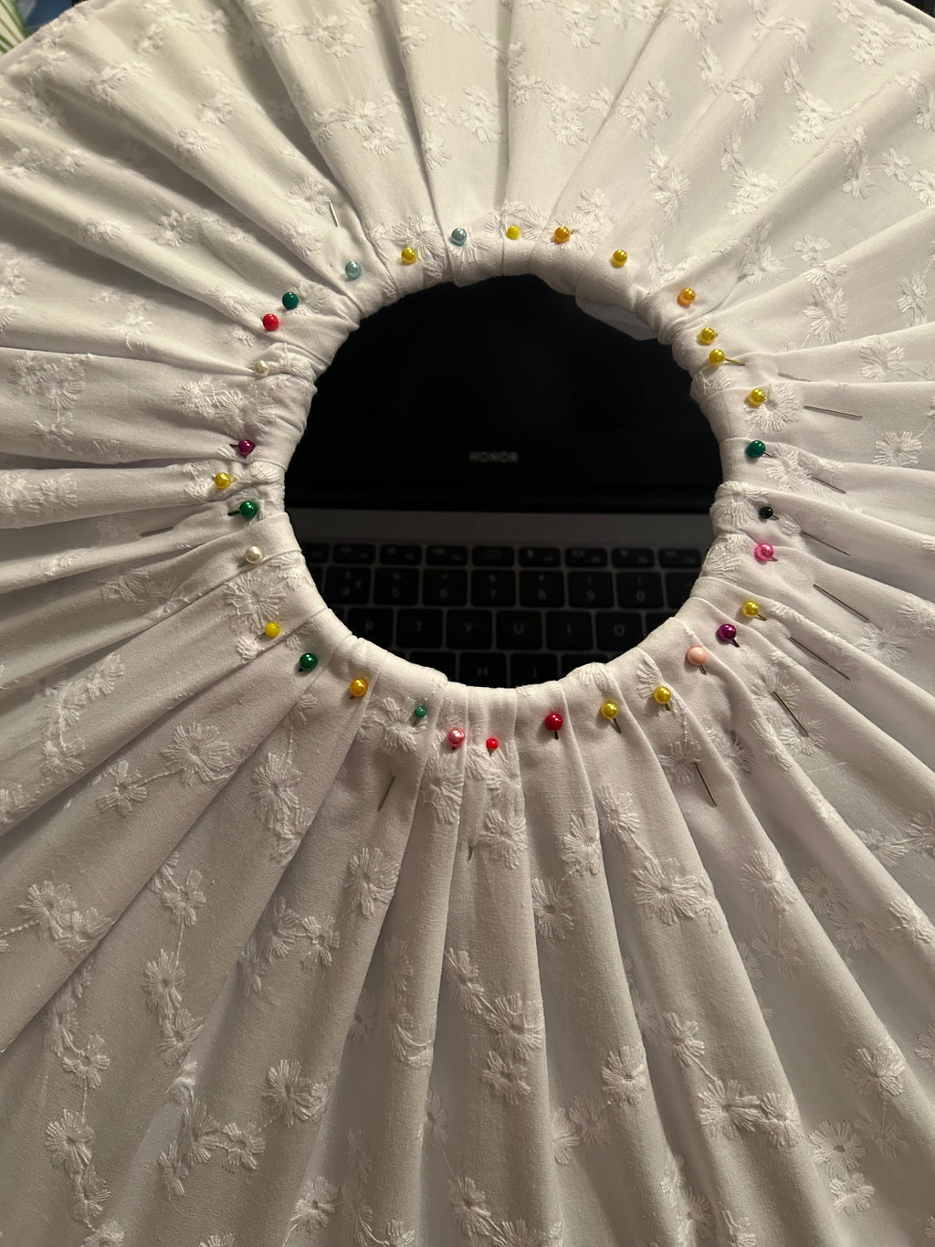 A close-up of the top view of a white broderie anglaise fabric shade with gathered radial pleats around a circular opening, secured with colourful sewing pins. A laptop is visible in the background through the opening. This is an 'in production' shot