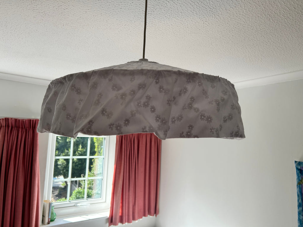 A fabric-covered dome-shaped pendant light shade with a white broderie anglaise floral pattern, hanging from a cord in a room with white walls, a textured ceiling, and pink curtains.