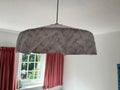 A fabric-covered dome-shaped pendant light shade with a white broderie anglaise floral pattern, hanging from a cord in a room with white walls, a textured ceiling, and pink curtains.
