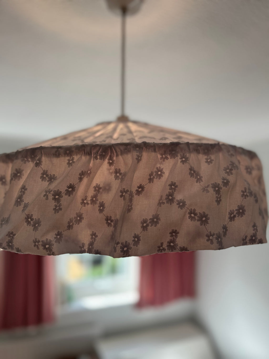 A vintage-style cottage core broderie anglaise pendant lampshade made of sheer white fabric with the floral pattern more prominent as the lightbulb is on, shown hanging in a softly blurred room.