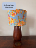 A table lamp with a glossy dark brown wooden base and a cylindrical fabric shade featuring a colourful abstract floral pattern in peach yellow and pale blue on a light blue background. A light blue text bubble in the top left reads, 'My lining is sky blue linen'.