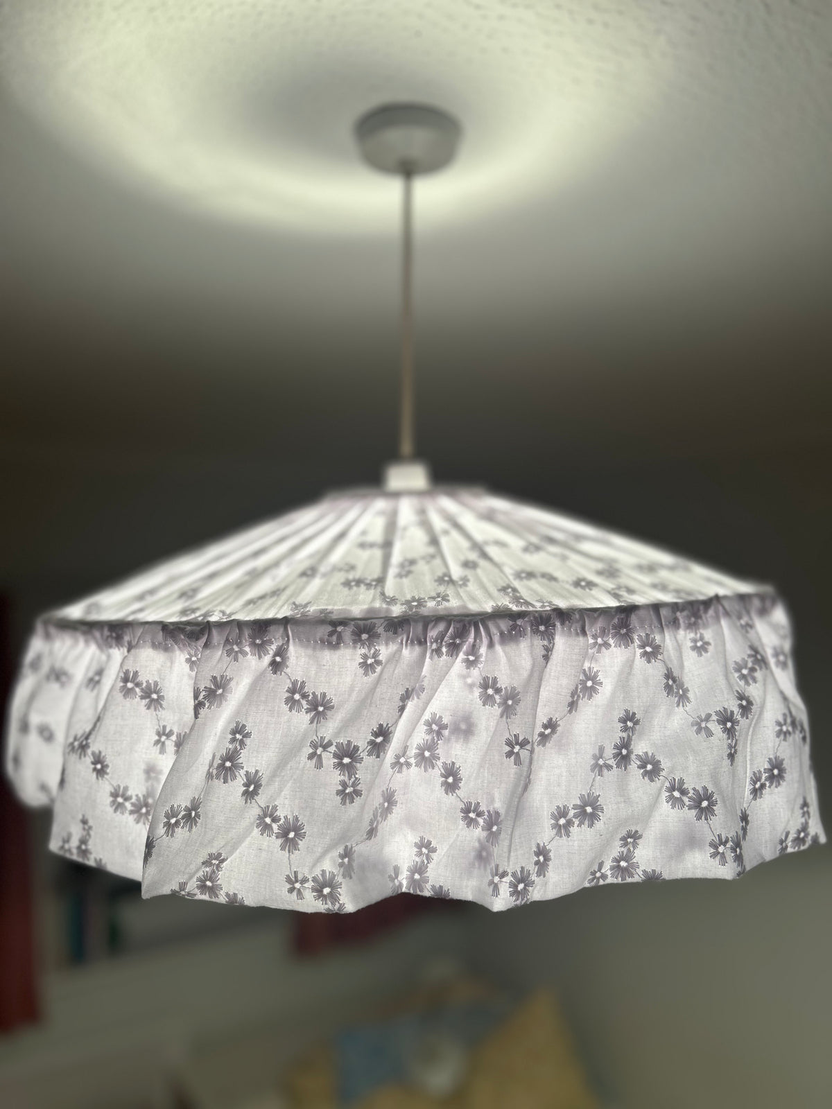 A fabric white broderie anglaise pendant lamp shade with a delicate floral pattern, hanging from a ceiling in a home interior.