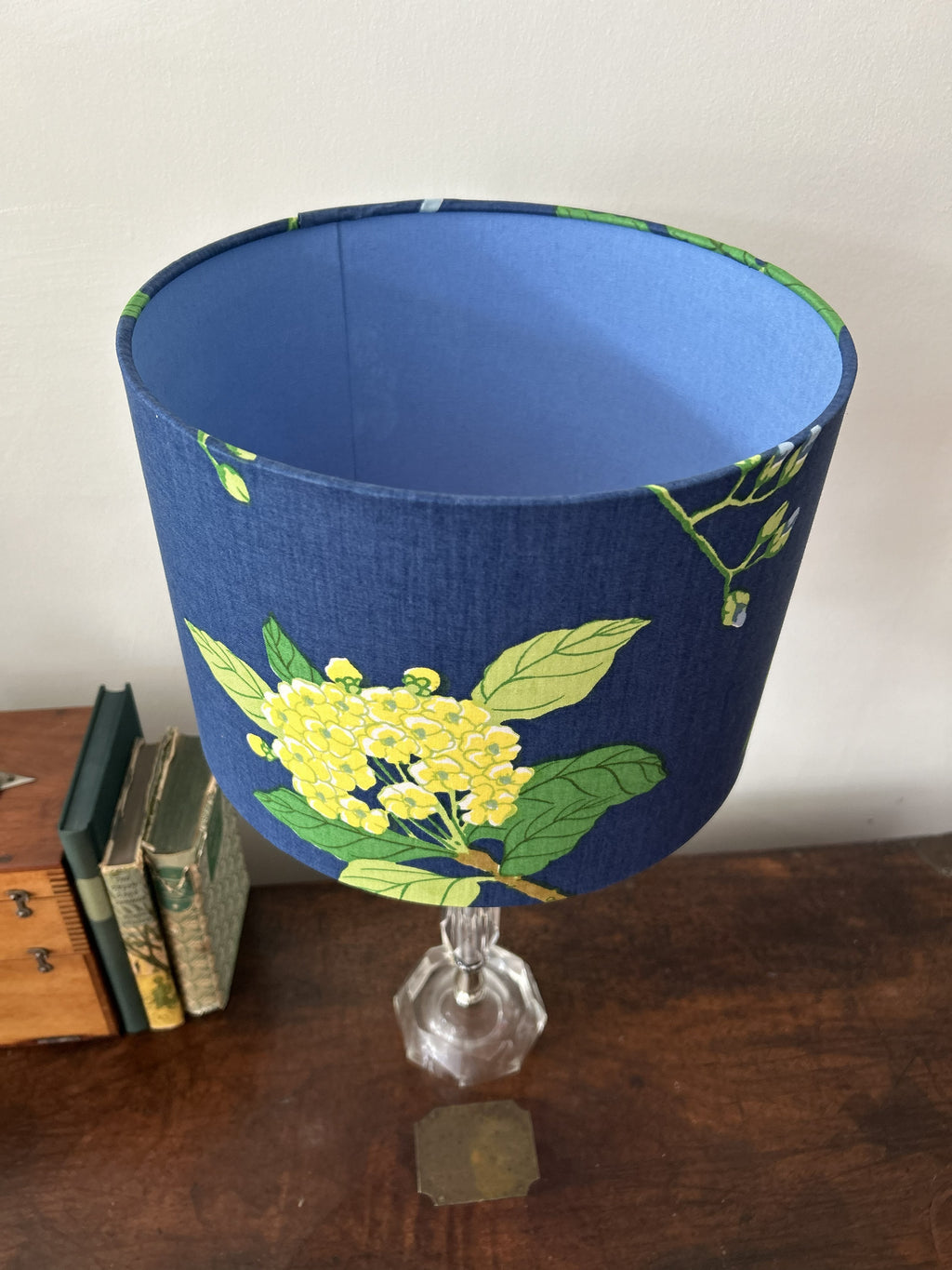 A fabric lampshade with a blue base and a yellow floral print, mounted on a clear faceted glass stem lamp. The lamp is set on a dark wooden table next to some vintage books against a plain off-white wall.